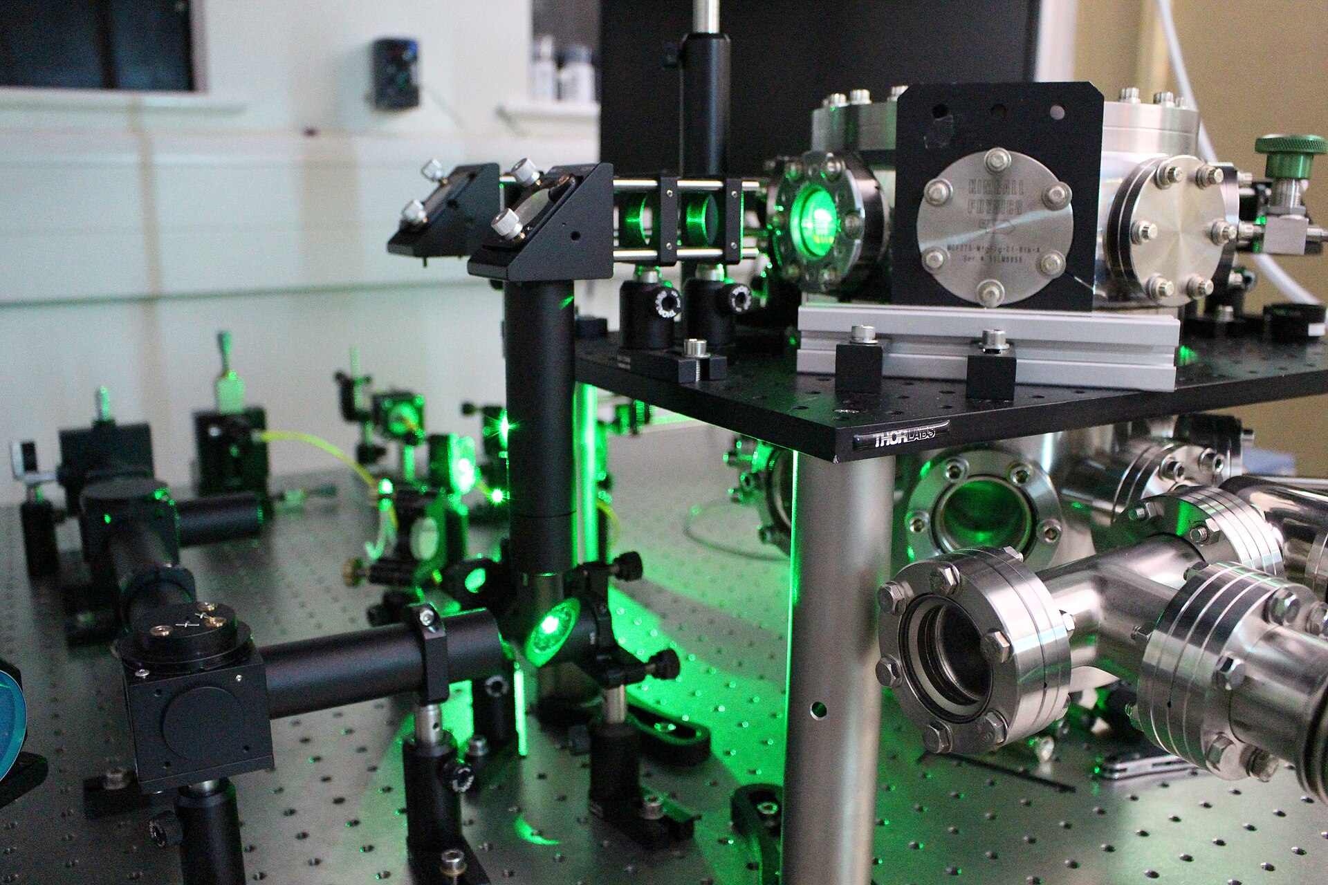 Banc optique de laboratoire avec miroirs, lentilles et chambre à vide traversés par une lumière laser verte