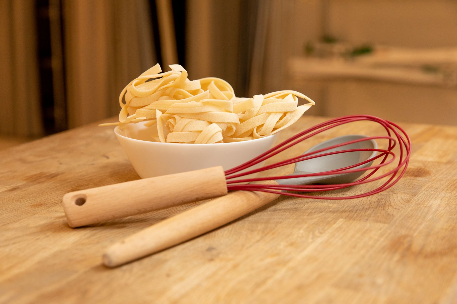 Bol blanc de tagliatelles sèches sur une table en bois, avec un fouet à fils rouges et une cuillère à manche en bois au premier plan.